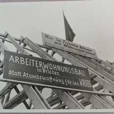 Schwarz-Weiß-Bild mit Dach und Schildern "ARBEITERWOHNUNGSBAU in Frieden statt Atombewaffnung für den Krieg"