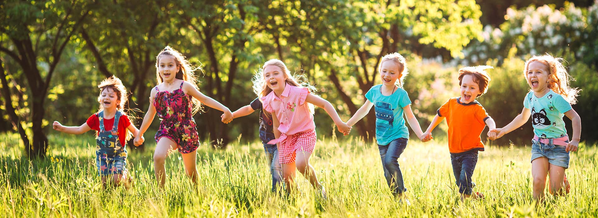 Kinder laufen Hand in Hand lachend auf einem Feld mit Bäumen im Hintergrund und Sonnenschein